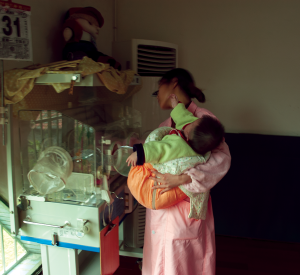 A caregiver holds an older baby in one arm while also tending a newborn in special care at an SWI in China.