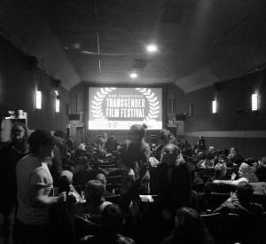 A black and white image of a crowd in a room with "San Fransisco Transgender Film Festival" projected on a screen