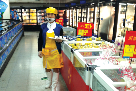 A migrant woman sells yogurt in a Beijing Walmart. She holds out free samples on her tray and wears a microphone over her face mask to announce the product and price to customers / photo by Eileen Otis.
