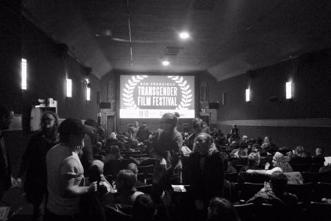 A black and white image of a crowd in a room with "San Fransisco Transgender Film Festival" projected on a screen