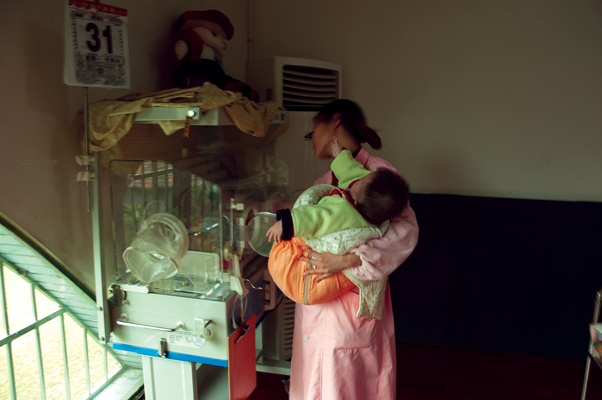 A caregiver holds an older baby in one arm while also tending a newborn in special care at an SWI in China.