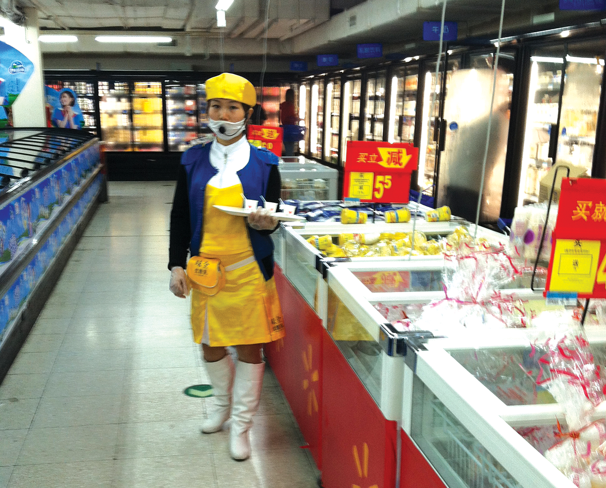 A migrant woman sells yogurt in a Beijing Walmart. She holds out free samples on her tray and wears a microphone over her face mask to announce the product and price to customers / photo by Eileen Otis.