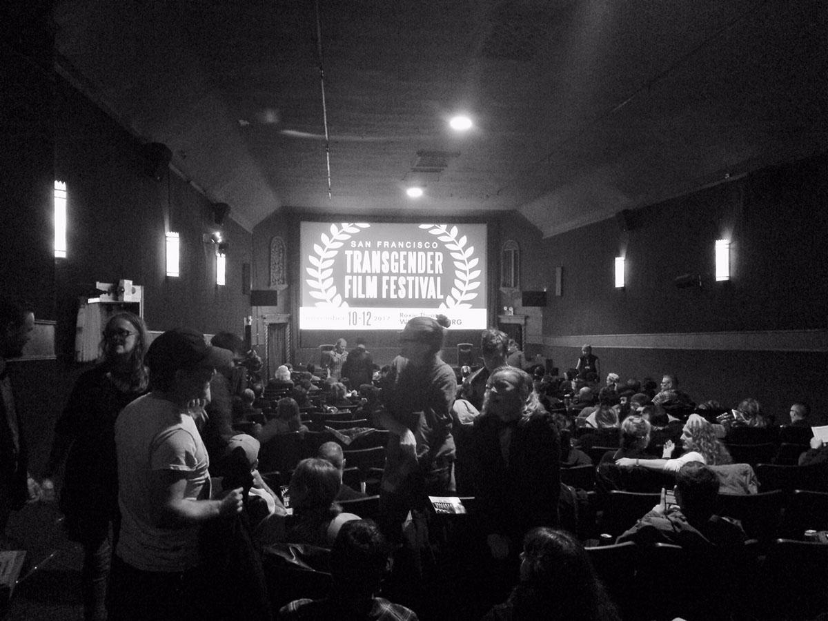 A black and white image of a crowd in a room with "San Fransisco Transgender Film Festival" projected on a screen