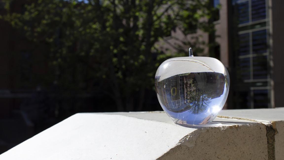 A crystal apple sits on a ledge in the sunshine reflecting Lillis Hall at the University of Oregon.